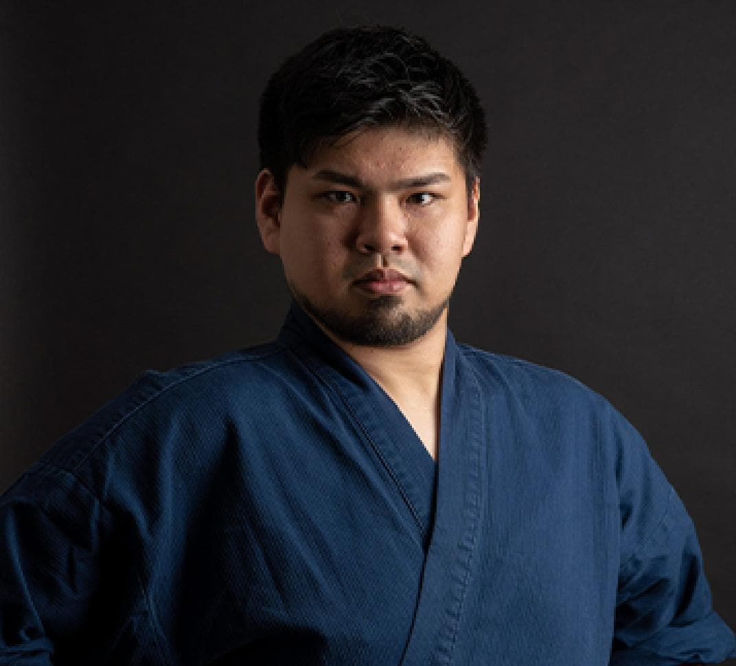 Japanese man in blue robe looks centered in front of back blackground.