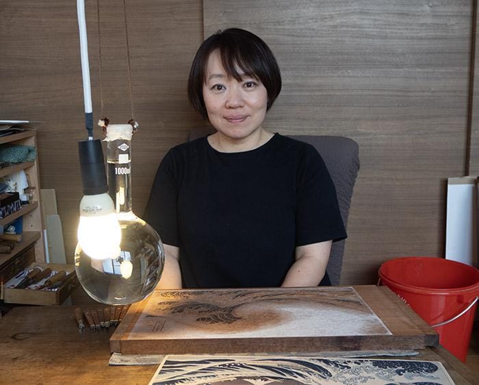 Japanese woman with mid-length dark brown hair sits in front of a woodblock to be carved of Hokusai's Great Wave.